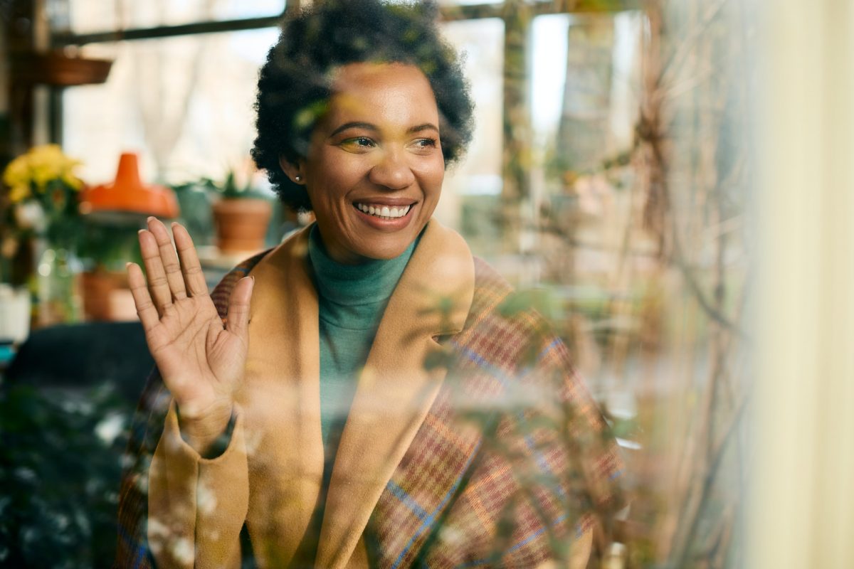 Happy black woman waving to someone through the window of coffee shop.